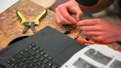 Hænder der forbinder ledninger på elektronik på et værkstedsbord med værktøj og en bærbar computer ved siden af
